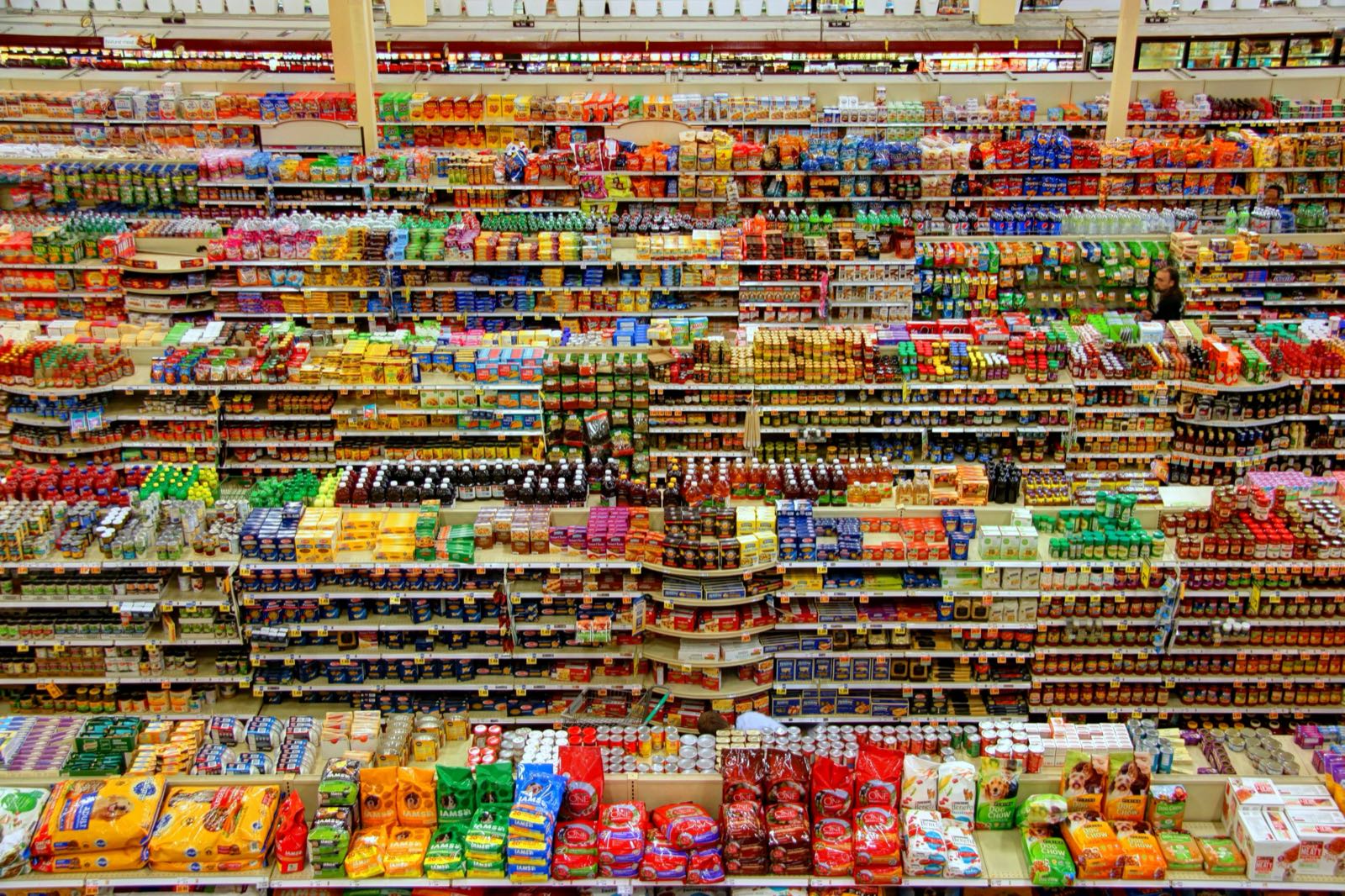 Brightly lit grocery store aisle with colorful produce and shoppers browsing